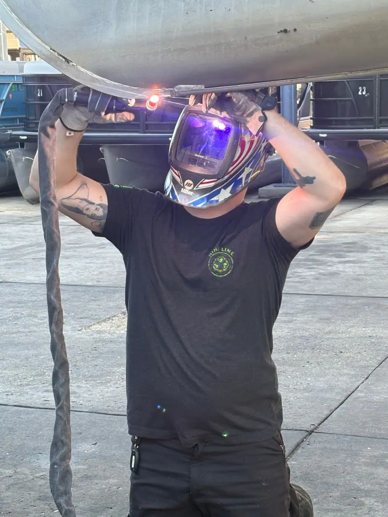 Welder kneeling under a pontoon tube at a boat yard with rental pontoons lined up behind